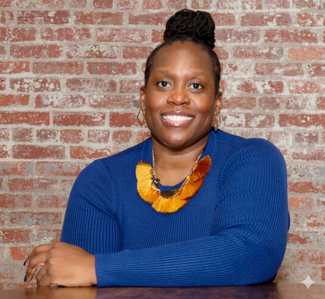 Portrait of a smiling woman with braided hair in a bun, wearing a blue sweater, gold earrings, and a colorful necklace, sitting in front of a brick wall.