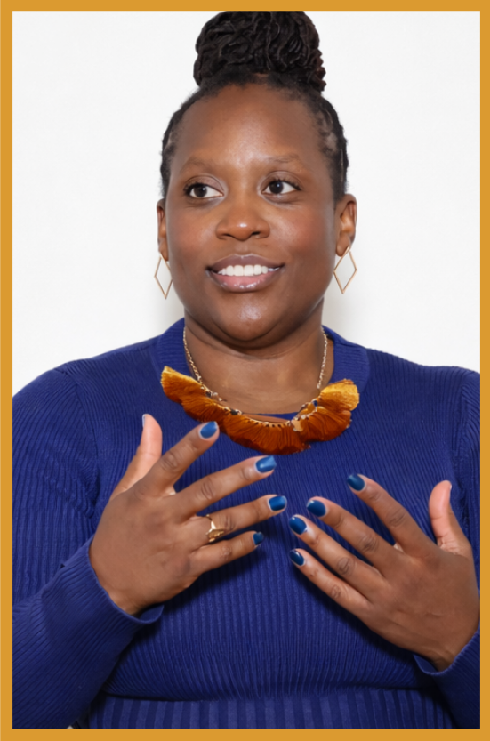 A woman with braided hair in a bun, wearing a blue sweater, gold earrings, and a large brown statement necklace, smiling and touching her chest with her right hand.