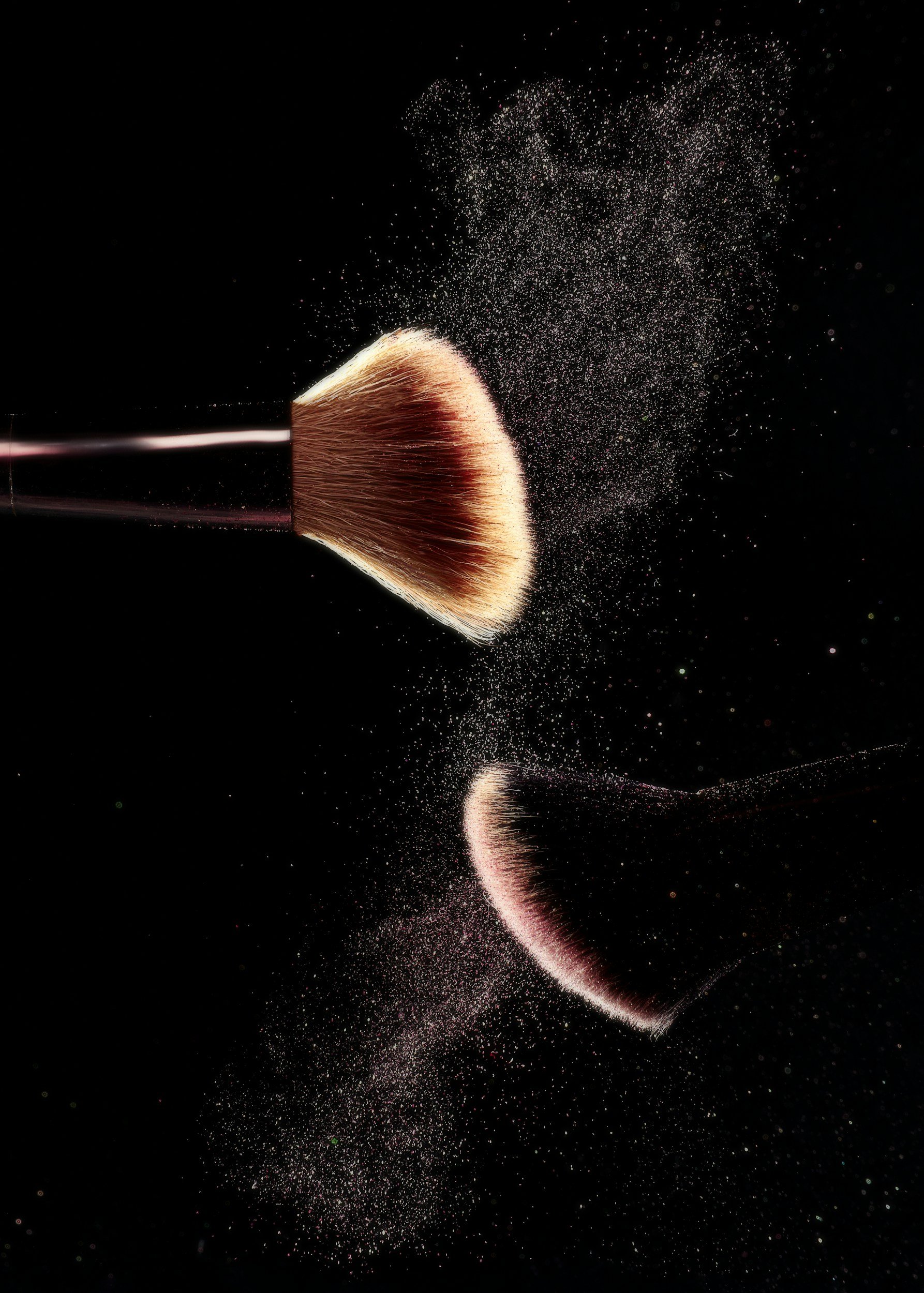 A makeup brush releasing powder or blush, which disperses into the air and reflects on a shiny black surface.
