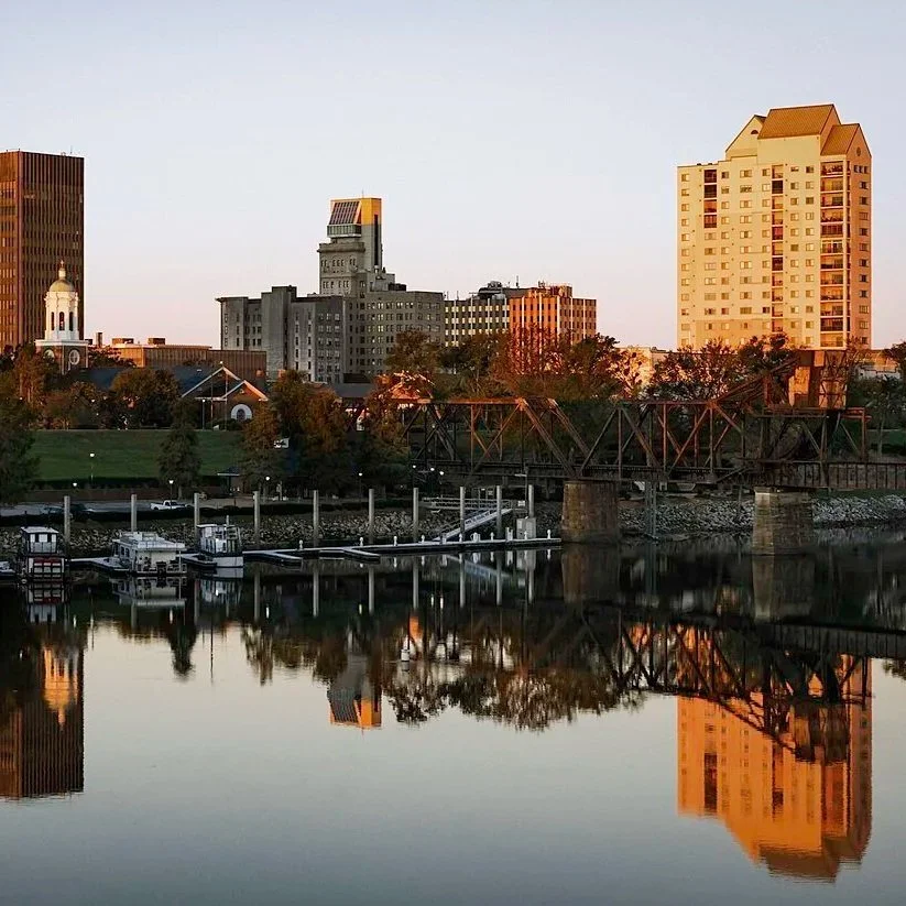 City skyline with tall buildings, a church with a white dome, trees, a bridge, and boats docked along a calm river at sunset.