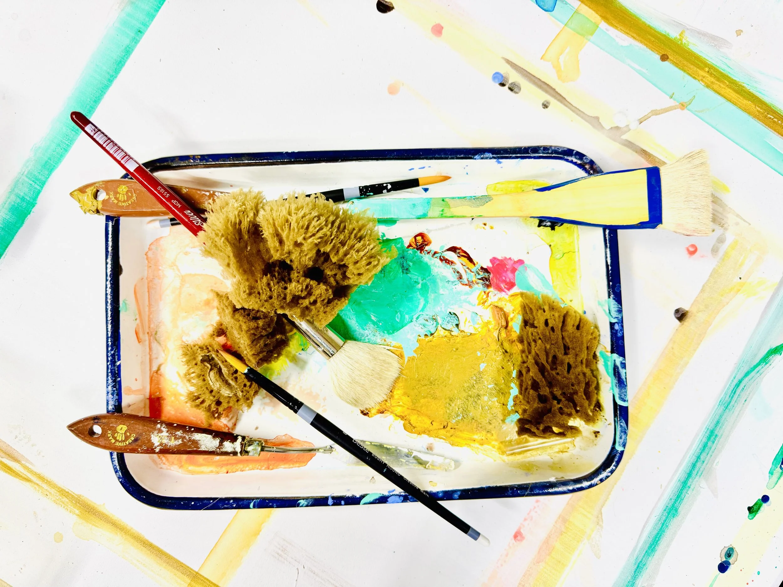 A small paint palette with various colors of paint, surrounded by paint brushes and markers, on a white surface with colorful paint splatters and streaks.