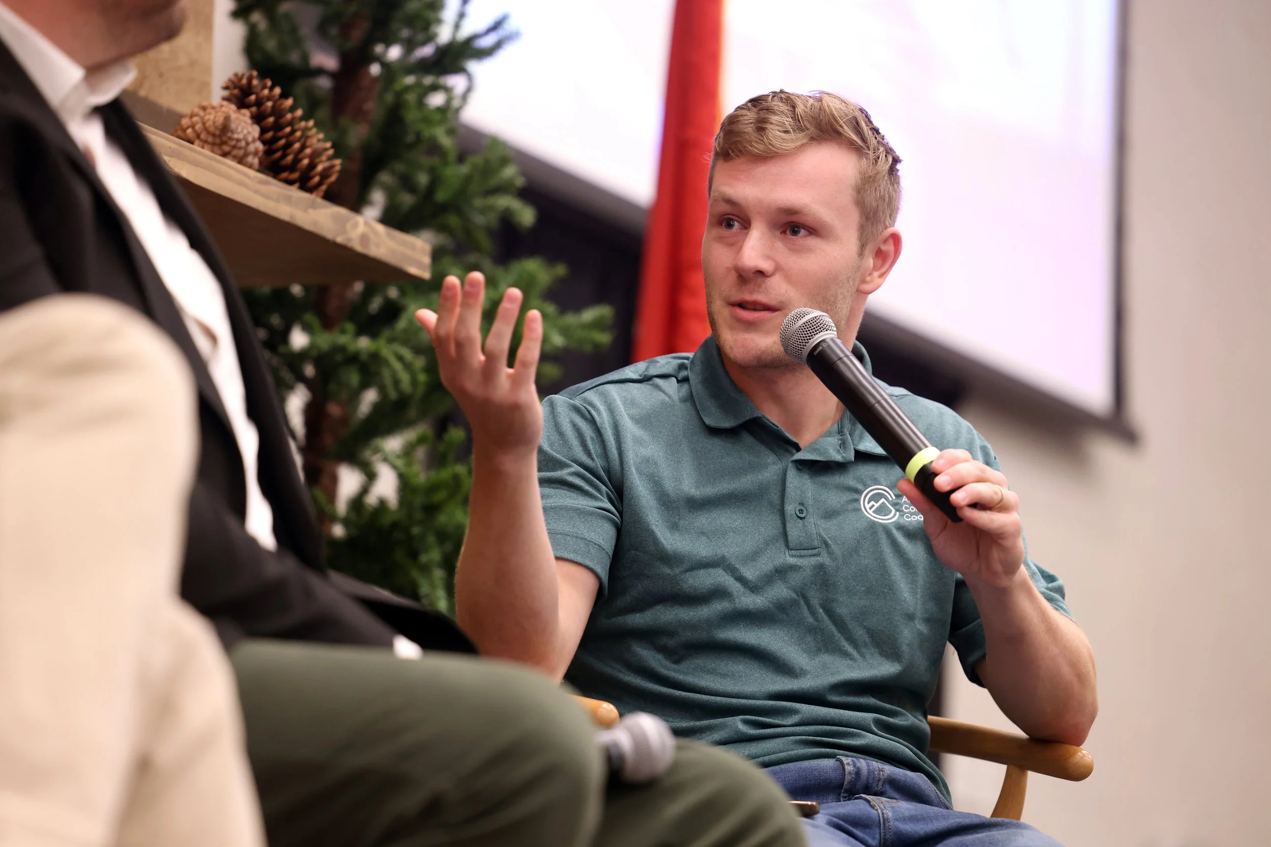 American Conservation Coalition (ACC) President Chris Barnard, sitting on a chair with a background decorated with pinecones and greenery, speaks into microphone during a panel conversation.