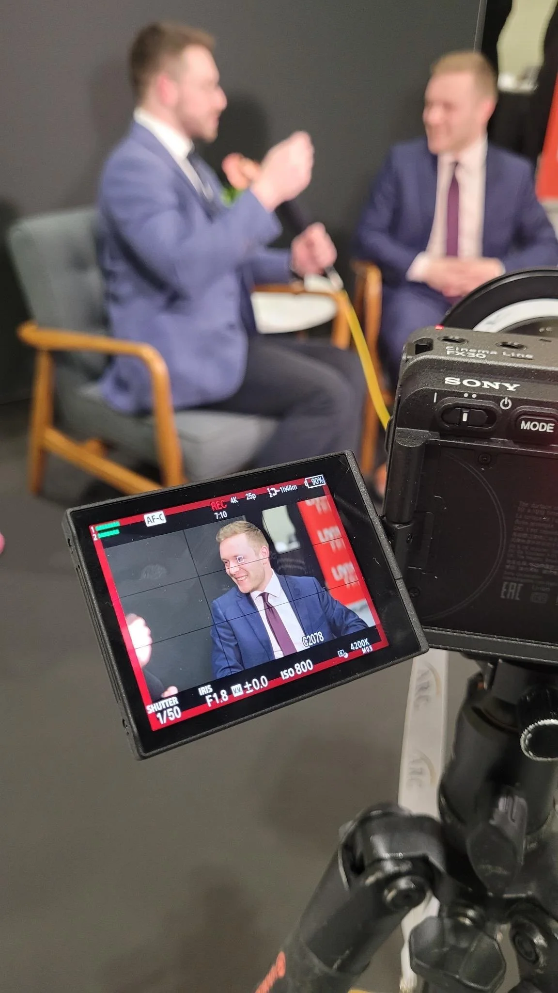 American Conservation Coalition (ACC) President Chris Barnard speaks into a microphone during an interview, sitting in a gray chair, with another man in a suit listening.
