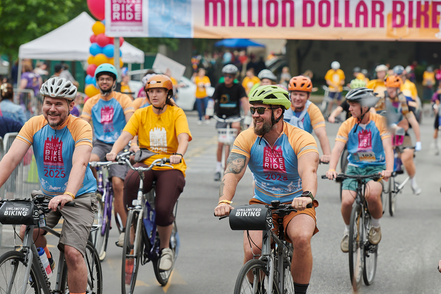People riding bicycles during a cycling event called Million Dollar Bike Ride 2025, wearing colorful t-shirts and helmets, with a banner overhead and a crowd in the background.