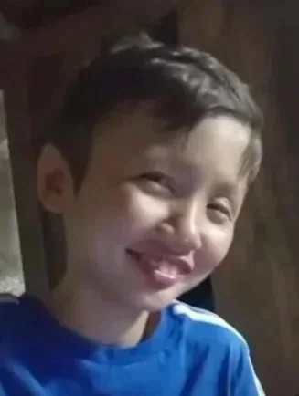 A smiling young boy with short dark hair, wearing a blue shirt in an indoor setting.