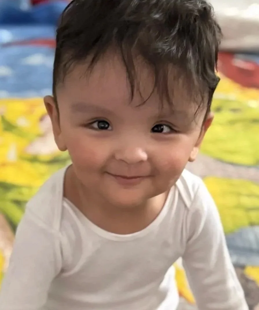 A smiling young child with dark hair and large eyes, wearing a white shirt, on a colorful patterned background.