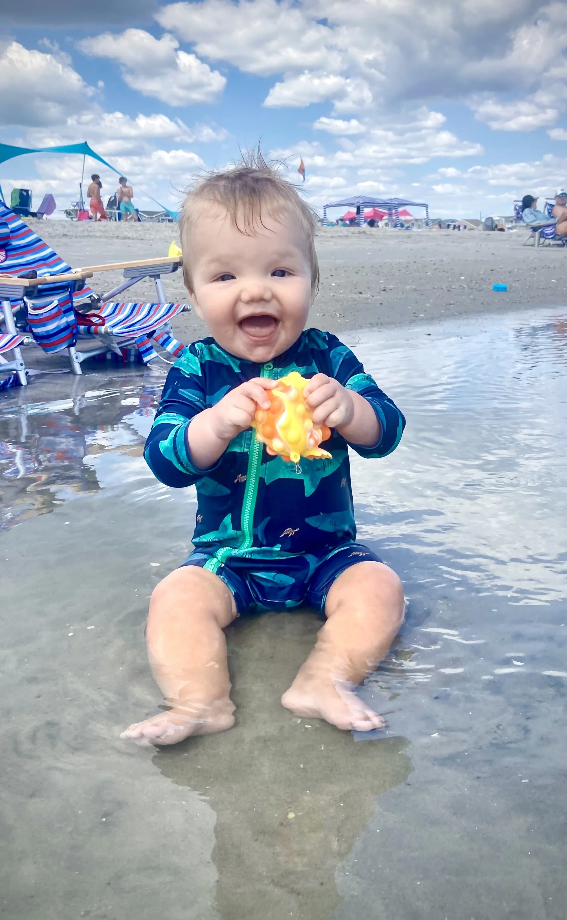 A smiling baby sitting in shallow water at the beach, holding a yellow toy, with beach chairs and umbrellas, and other people in the background under a partly cloudy sky.