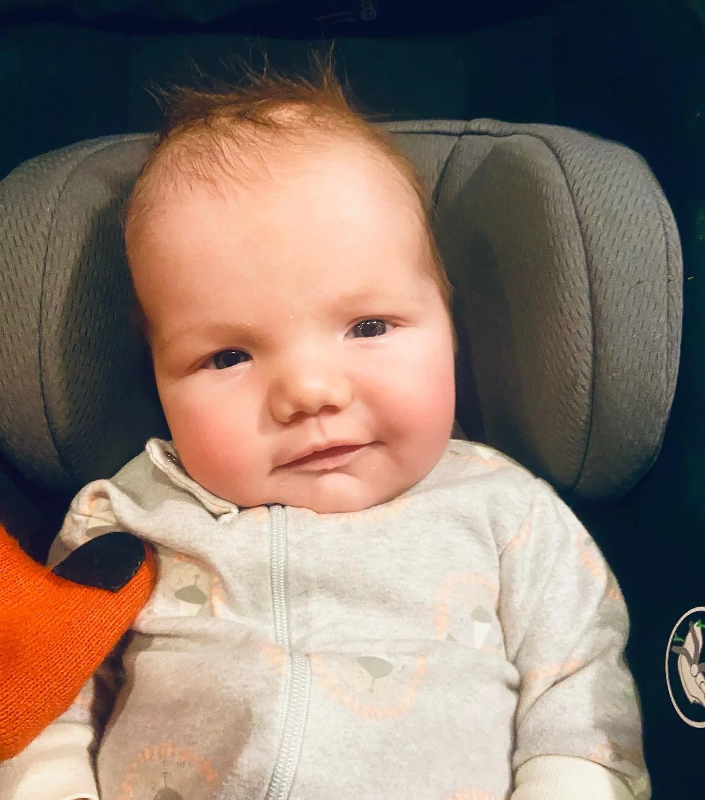 Close-up of a smiling baby with light skin and brown hair, sitting in a car seat.
