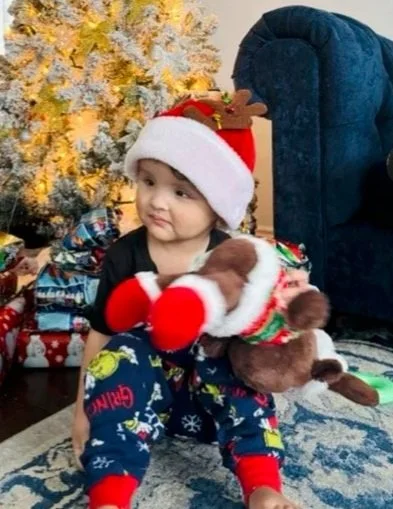 A young child wearing a Santa hat and Christmas pajamas sitting on the floor and holding a plush reindeer toy, with a decorated Christmas tree and wrapped presents in the background.