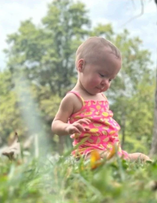 A young girl in a pink and yellow outfit sitting outdoors among green grass and flowers.