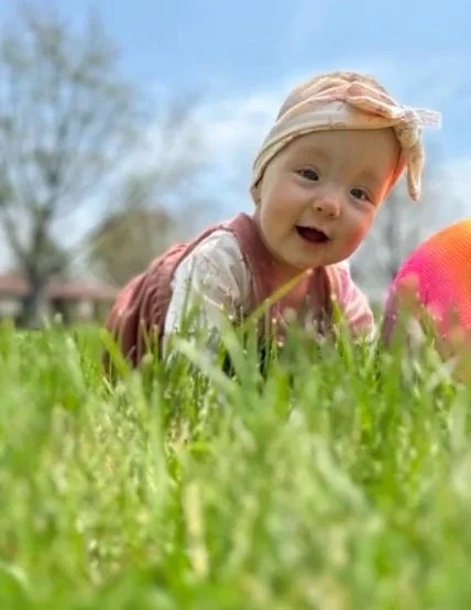 A baby girl crawling on grass outdoors, wearing a light-colored headband and pink vest, smiling at the camera.