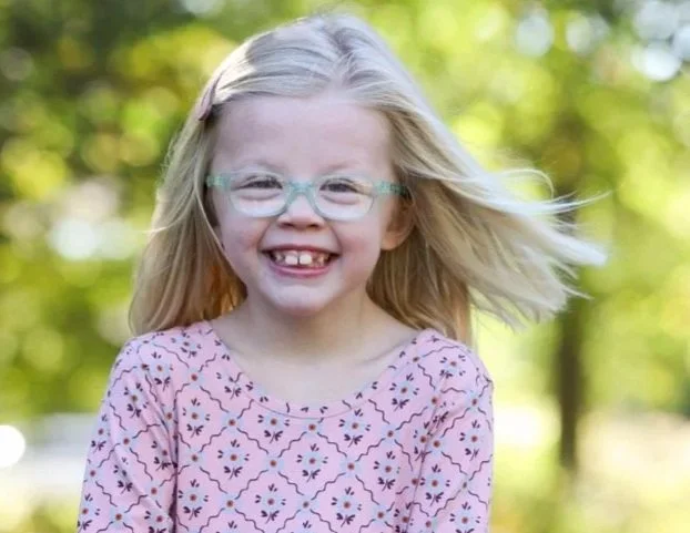 A young girl with blonde hair, glasses, and a pink patterned dress is smiling outdoors with a blurred green background.