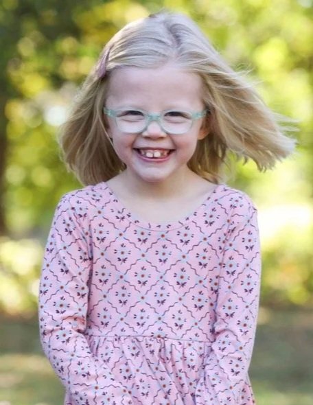 A young girl with blonde hair, glasses, and a pink patterned dress smiling outdoors.