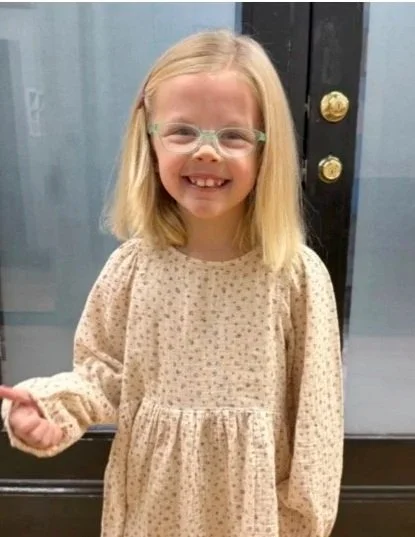 A young girl with blonde hair, glasses, and a beige polka-dot dress standing in front of a door, smiling at the camera.