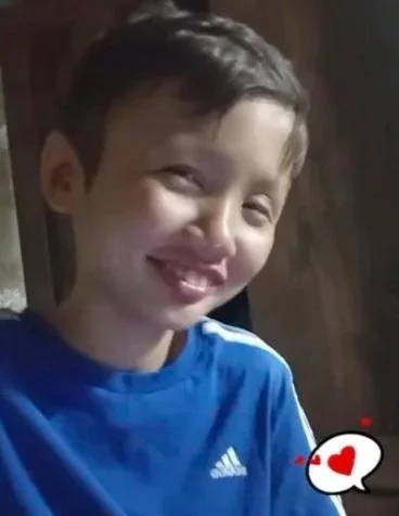 Smiling young boy with short dark hair wearing a blue Adidas shirt, standing in front of a wooden background.