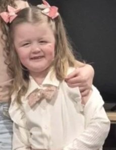 A young girl with curly hair tied with pink bows, wearing a white shirt, smiling and holding a small plush toy.