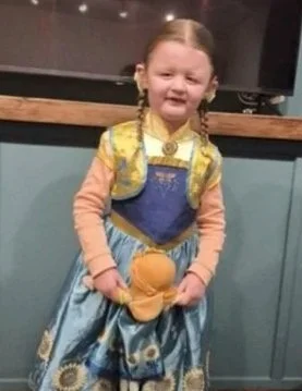 Young girl dressed as a princess, holding a plush toy, standing indoors.
