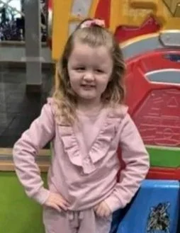 A young girl with long curly blonde hair wearing a pink outfit with ruffles, standing outdoors with her hands on her hips, smiling in front of colorful playground equipment.