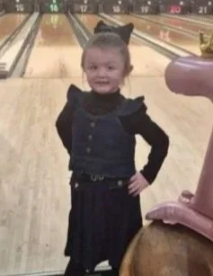 Young girl with hair pulled back, wearing a black dress, posing at a bowling alley.