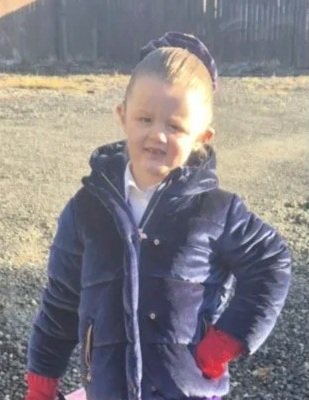 Child in a navy puffer jacket and red gloves standing outdoors with hands on hips, smiling, with a wooden fence in the background.