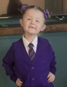 Young girl with pigtails smiling, dressed in a purple school uniform with a white shirt and striped tie.