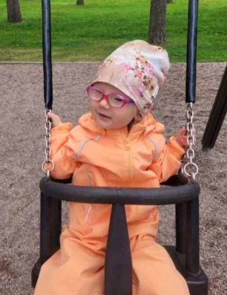 A young girl with glasses and a floral hat sitting on a swing at a park.