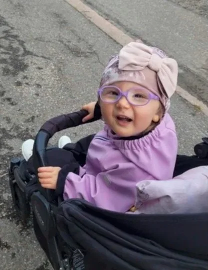 Smiling young girl with glasses and a bow headband sitting in a stroller on a paved street.