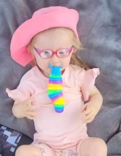 Young girl wearing pink glasses and a pink hat, sitting on a gray couch, with a rainbow-colored toy in her mouth.