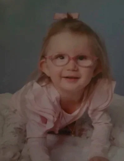A young girl with glasses and long blonde hair, wearing a pink bow, lying on her stomach and smiling, against a gray background.