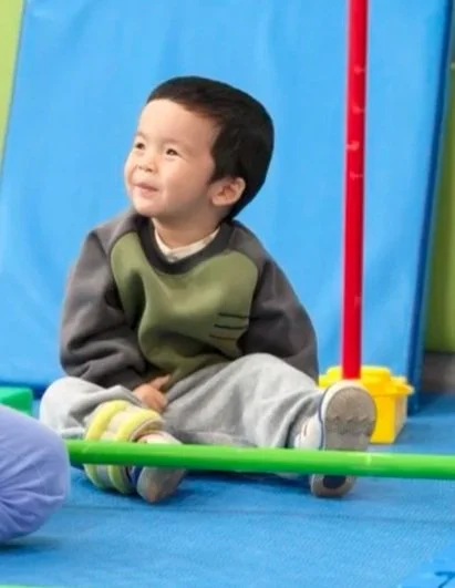 Young boy sitting on the floor in a play area, smiling and looking to the side, with colorful play equipment around him.