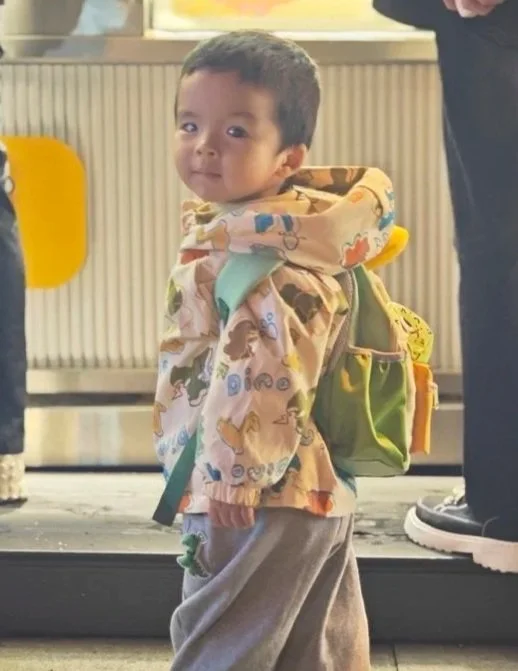 A young boy with short dark hair and a patterned jacket looking back at the camera, standing indoors with a yellow chair in the background.