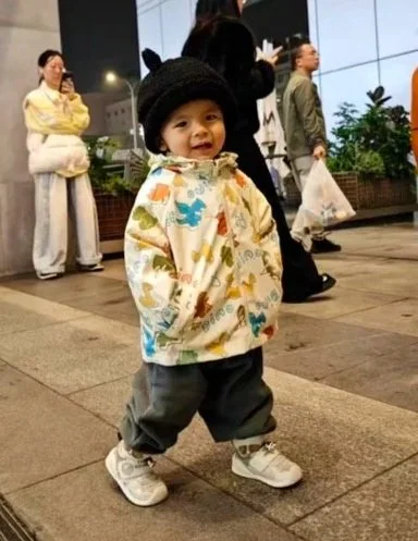 A young child standing outdoors at night, wearing a colorful jacket, gray pants, white sneakers, and a black hat with animal ears, with people walking in the background.