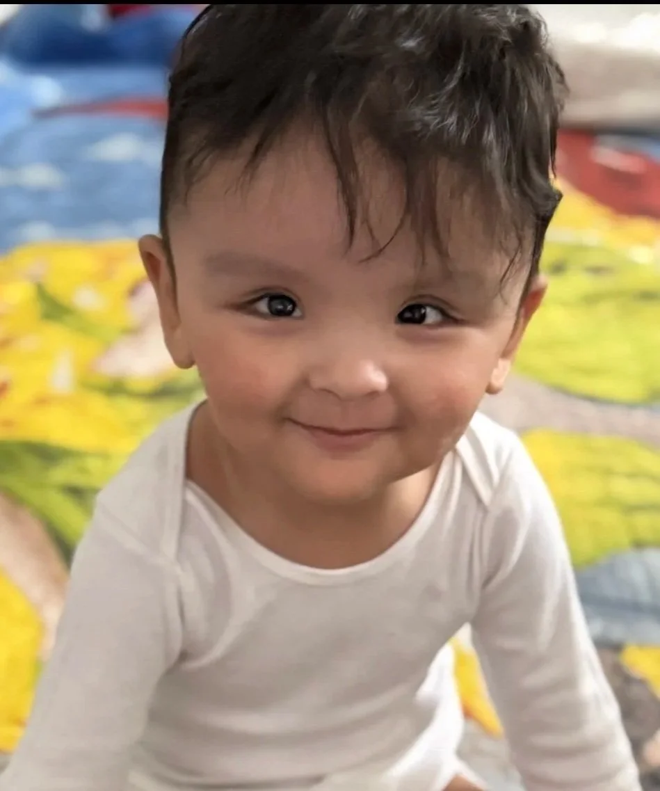 Close-up of a smiling young child with dark hair and brown eyes, wearing a white long-sleeve shirt, on a colorful patterned background.