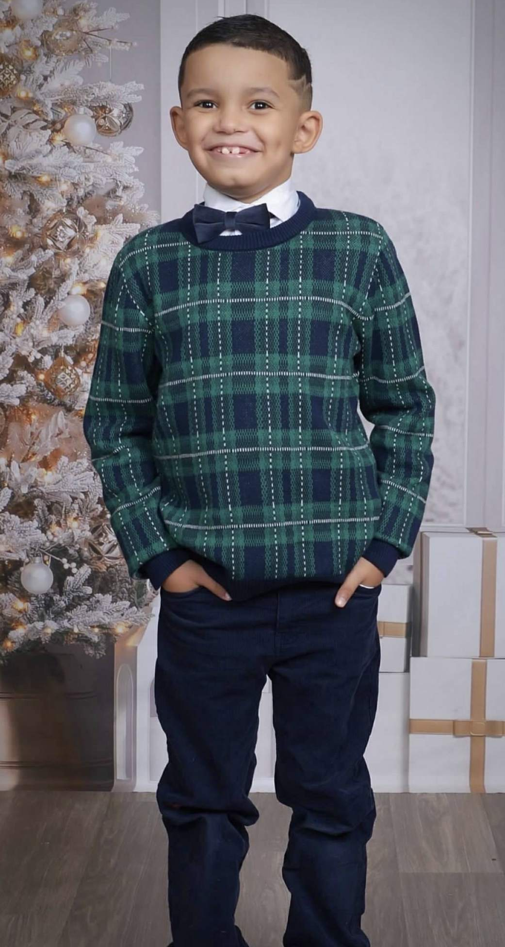 Young boy with a gap-tooth smile and short haircut, wearing a green plaid sweater, white collared shirt, black bow tie, and black pants, standing next to a decorated Christmas tree with white branches and ornaments.