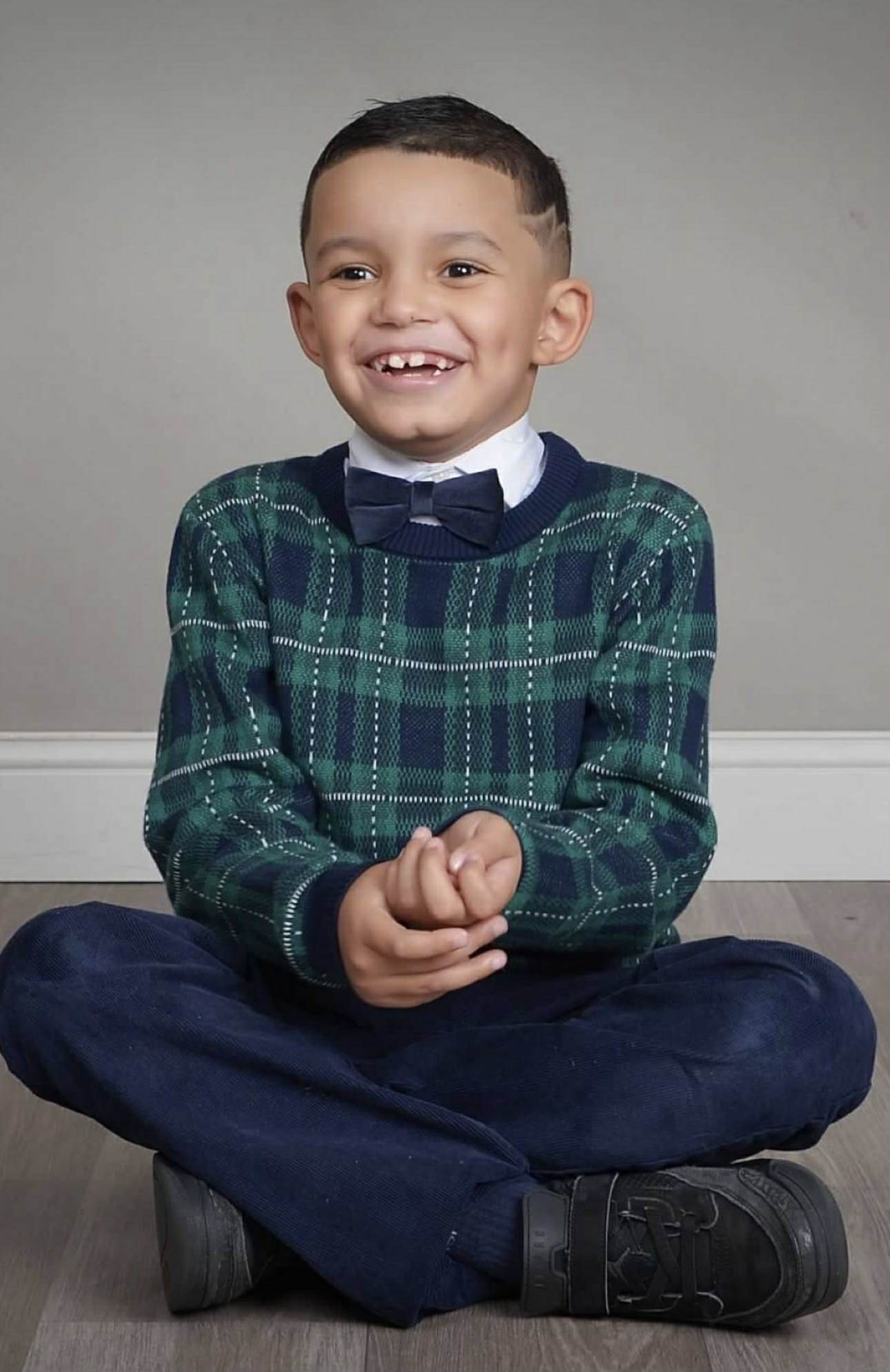 Smiling young boy with missing front teeth, sitting cross-legged on the floor, wearing a blue and green plaid sweater, white shirt with a bow tie, dark blue pants, and black shoes, against a plain gray wall background.