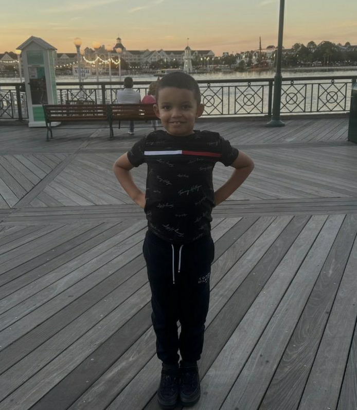 A young boy standing on a wooden pier with his hands on his hips, smiling at the camera during sunset. Behind him, there are benches, water, and a city skyline with buildings and a tower in the distance.