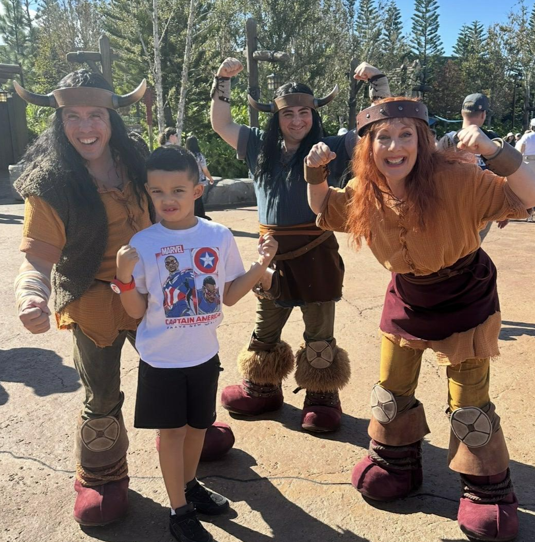 A boy stands with three women dressed as Viking characters at a theme park, all striking strong, victorious poses, outdoors on a sunny day.