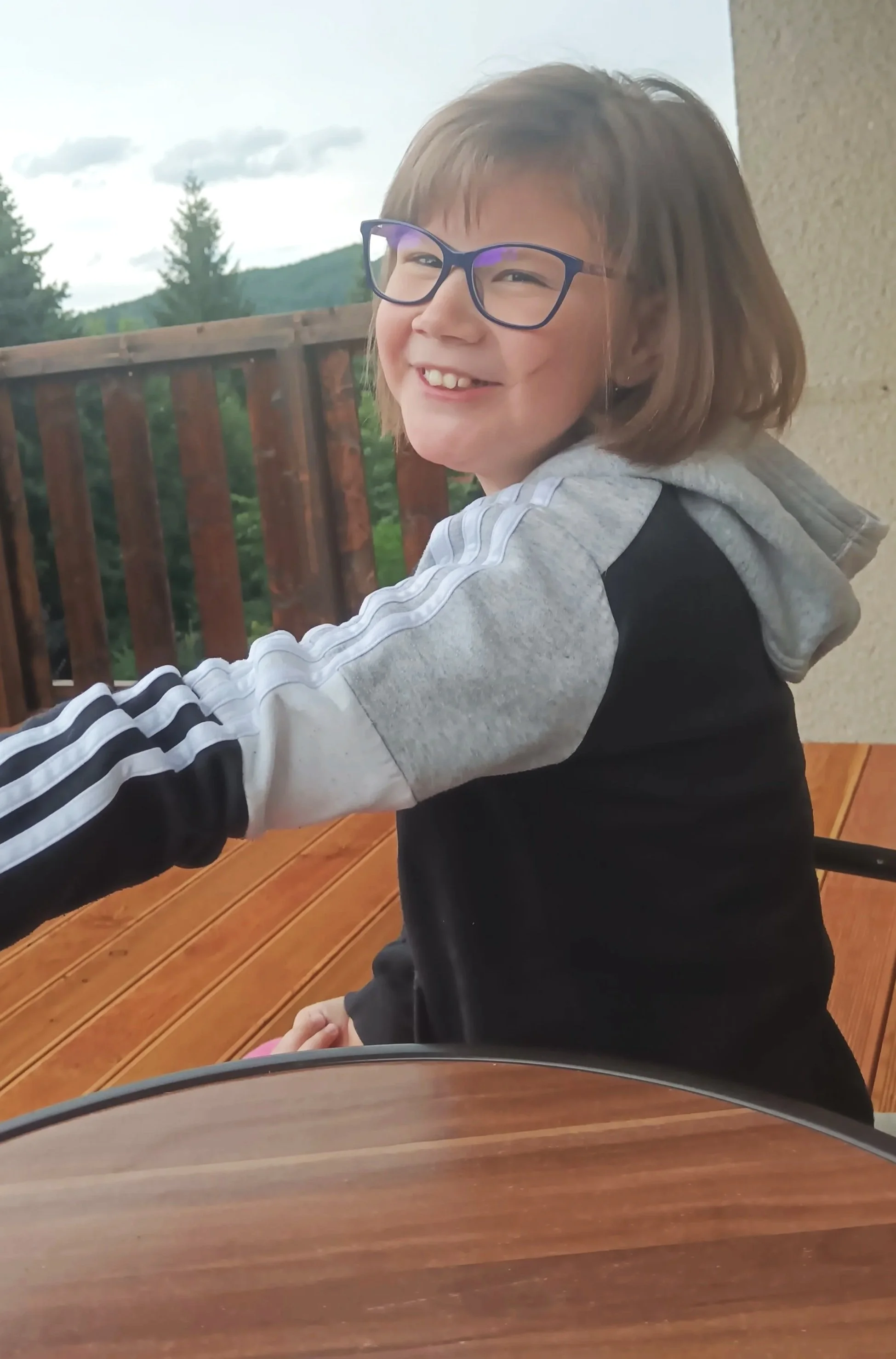A smiling young girl with glasses, wearing a gray and black hoodie with white and black striped sleeves, sitting outside on a wooden deck with a mountainous landscape and cloudy sky in the background.