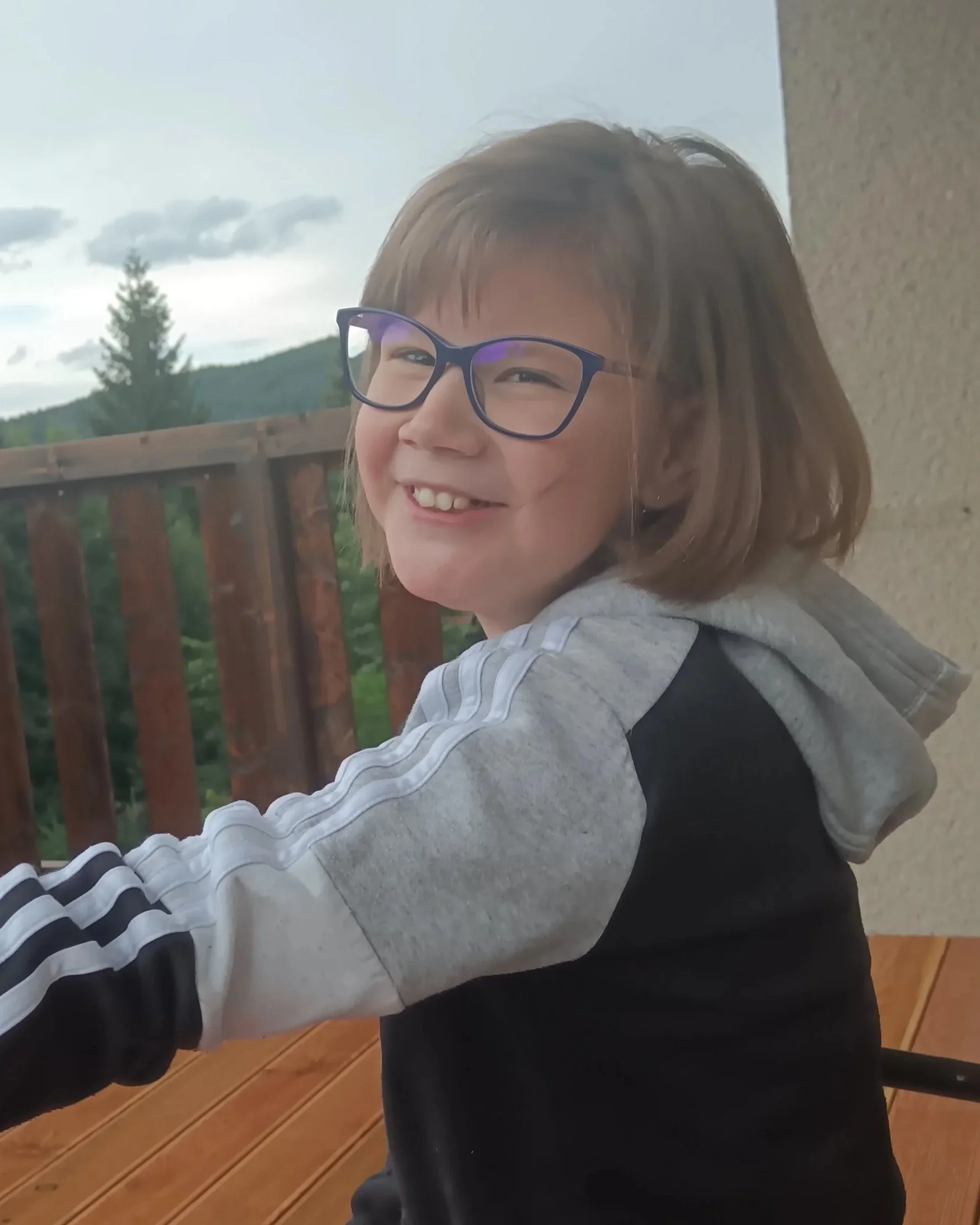 A smiling young girl with glasses and shoulder-length hair, wearing a gray and black hoodie, sitting on a porch with a wooden railing, in front of a scenic background of green trees and cloudy sky.