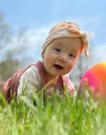 A smiling baby with a beige headband lying on grass outdoors, with a colorful ball nearby and a blue sky background.