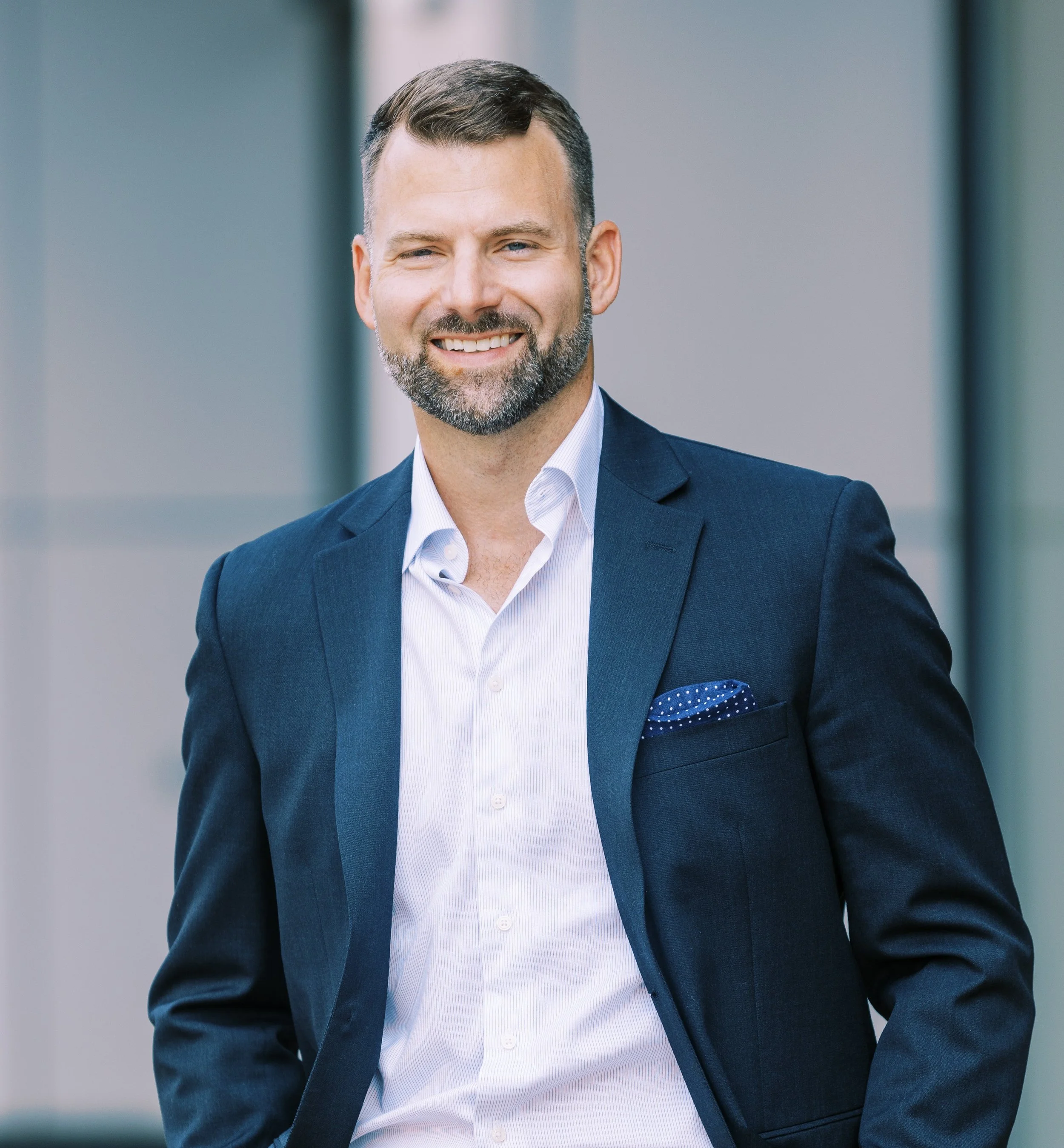 A man in a navy suit and white shirt smiling outdoors.