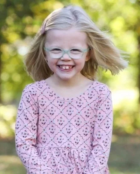 A young girl with blonde hair, wearing glasses and a pink patterned dress, smiling outdoors with blurred green trees in the background.