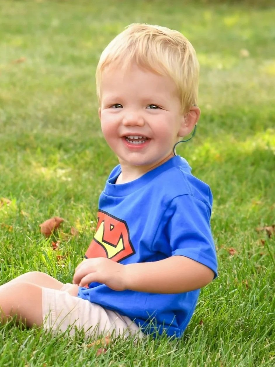 A young boy with blonde hair, wearing a blue Superman T-shirt, beige shorts, and a hearing aid, sitting on grass in a park and smiling at the camera.