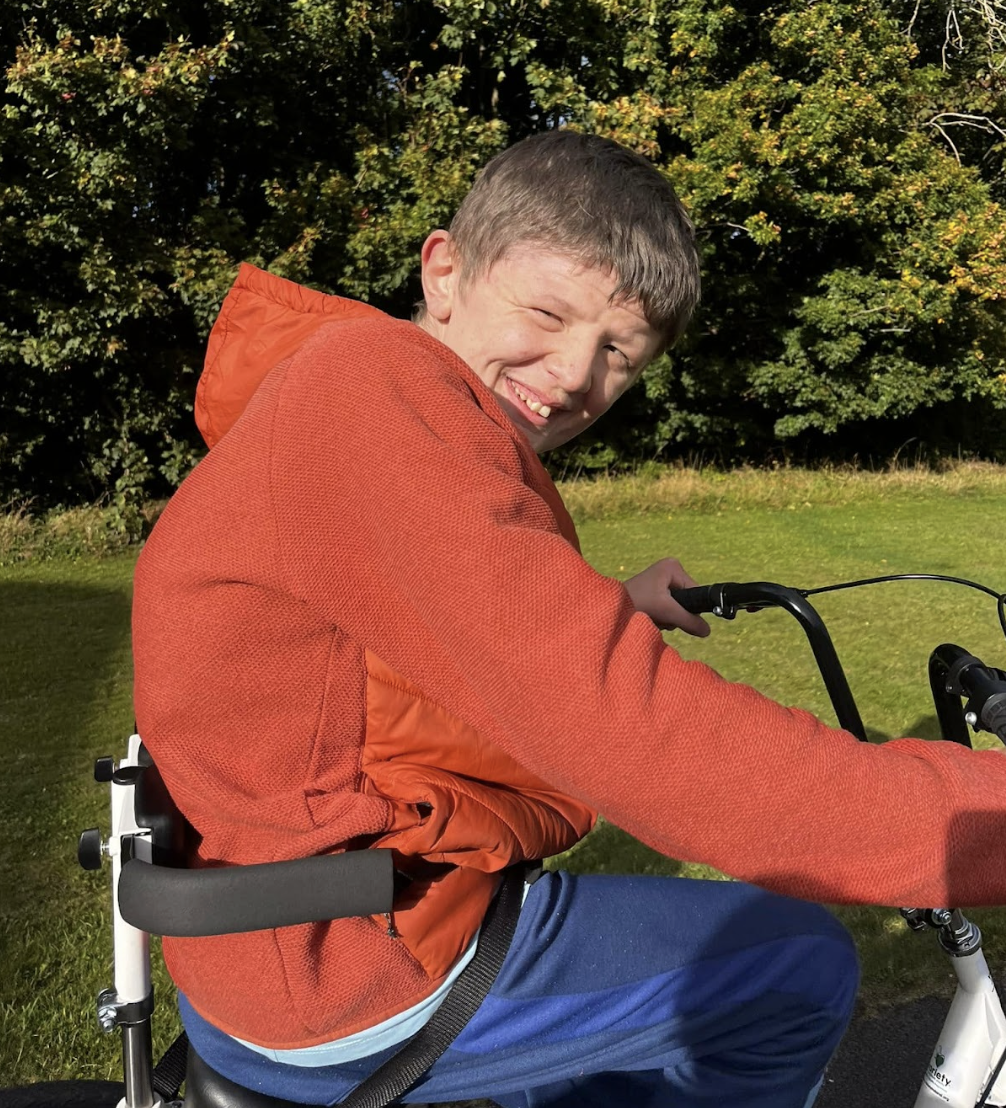A young boy smiling as he rides a bicycle outdoors on a sunny day, wearing an orange jacket and blue pants, with green trees in the background.
