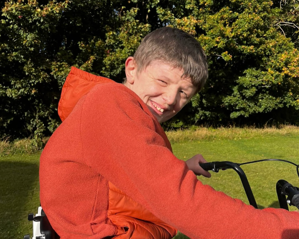 A young boy with short brown hair, smiling and winking, wearing an orange jacket, riding a bicycle in a grassy area with trees in the background.