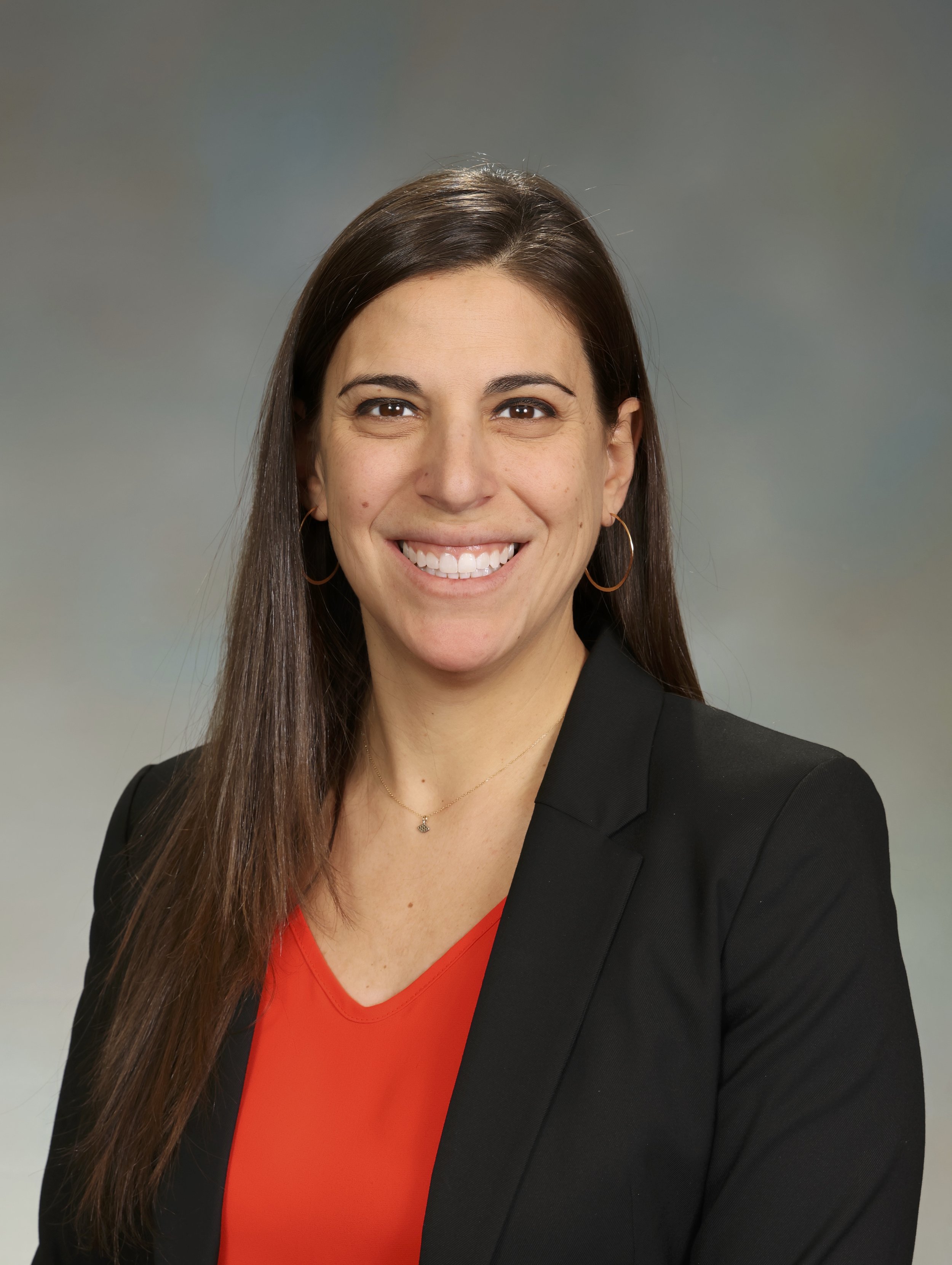 Portrait of a smiling woman with long dark hair, wearing a black blazer, red top, and gold jewelry, in front of a neutral background.