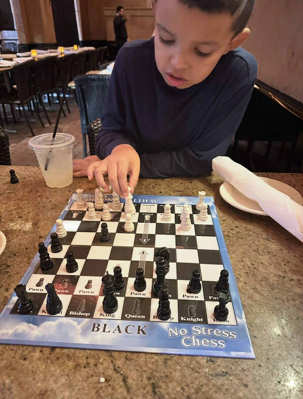 A young boy in a dark shirt playing chess at a restaurant table with a drink and empty plates nearby.