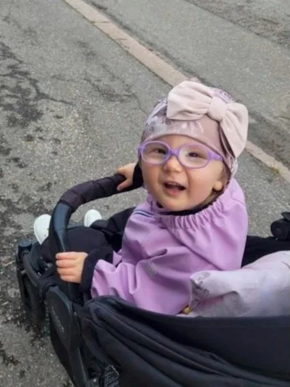 A young girl wearing glasses, a large bow headband, and a purple jacket sitting in a stroller on a sidewalk.