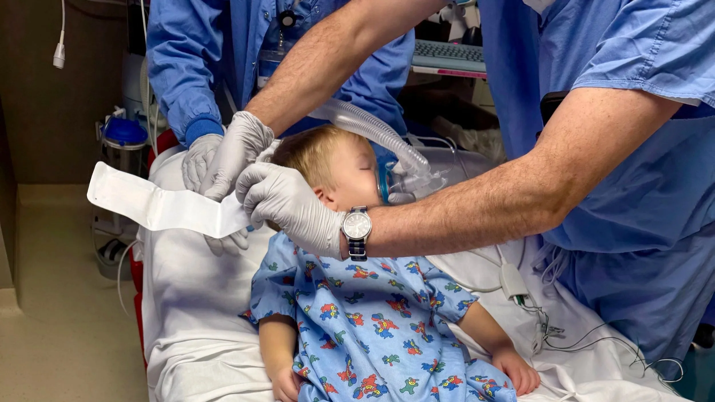 A young boy is lying on a hospital bed with his eyes closed, wearing an oxygen mask connected to a ventilator. Two healthcare professionals in blue scrubs are attending to him, one holding the mask in place while the other supports his head.