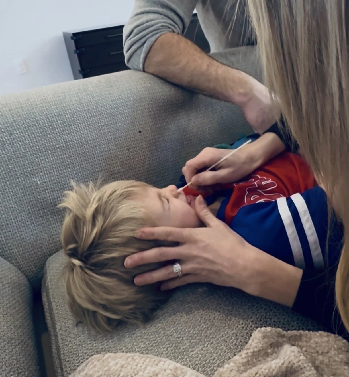 A young boy lying on a couch with a sippy straw in his mouth, being gently touched on the head by a woman wearing a wedding ring, as an adult male leans over from behind.